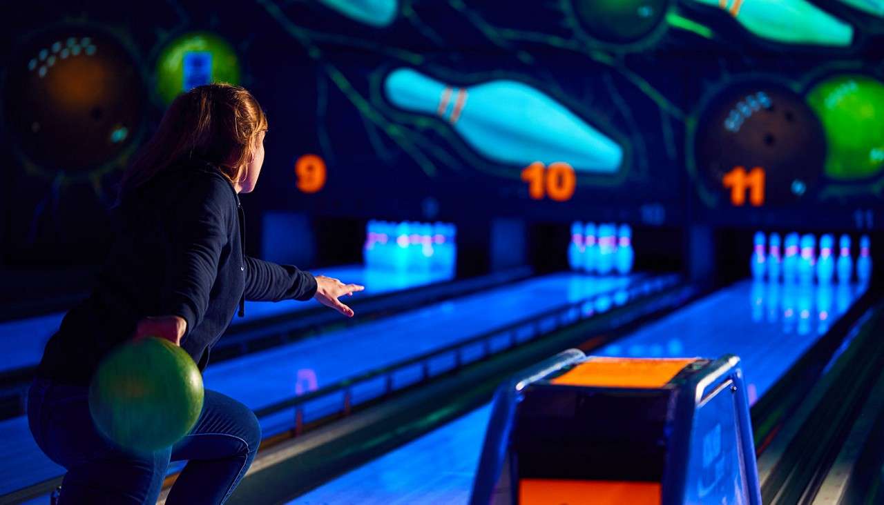 Bowler releases a green ball toward pins, aiming down a glowing blue lane in a neon-lit bowling alley. Text: 9, 10, 11.