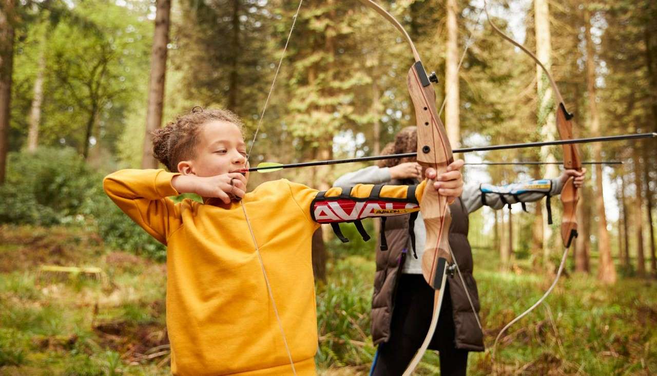 Child archer draws a traditional bow, aiming an arrow; another archer mirrors behind in a sunlit forest clearing. Text on armguard: AVA.