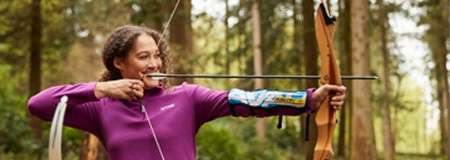 Archer draws a wooden recurve bow, arrow at full draw, wearing a purple top and armguard; standing sideways and aiming amid tall trees in a forest setting.
