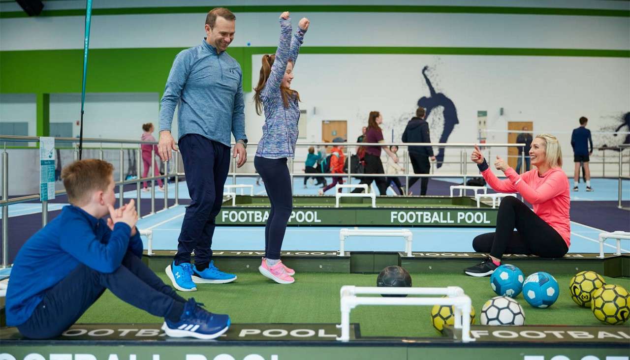 Family plays football pool; a girl celebrates with raised arms while a man smiles, a seated woman gives thumbs-up, and a boy watches. Indoor sports hall with green tables, soccer balls, and onlookers. Text: FOOTBALL POOL FOOTBALL POOL FOOTBALL POOL.
