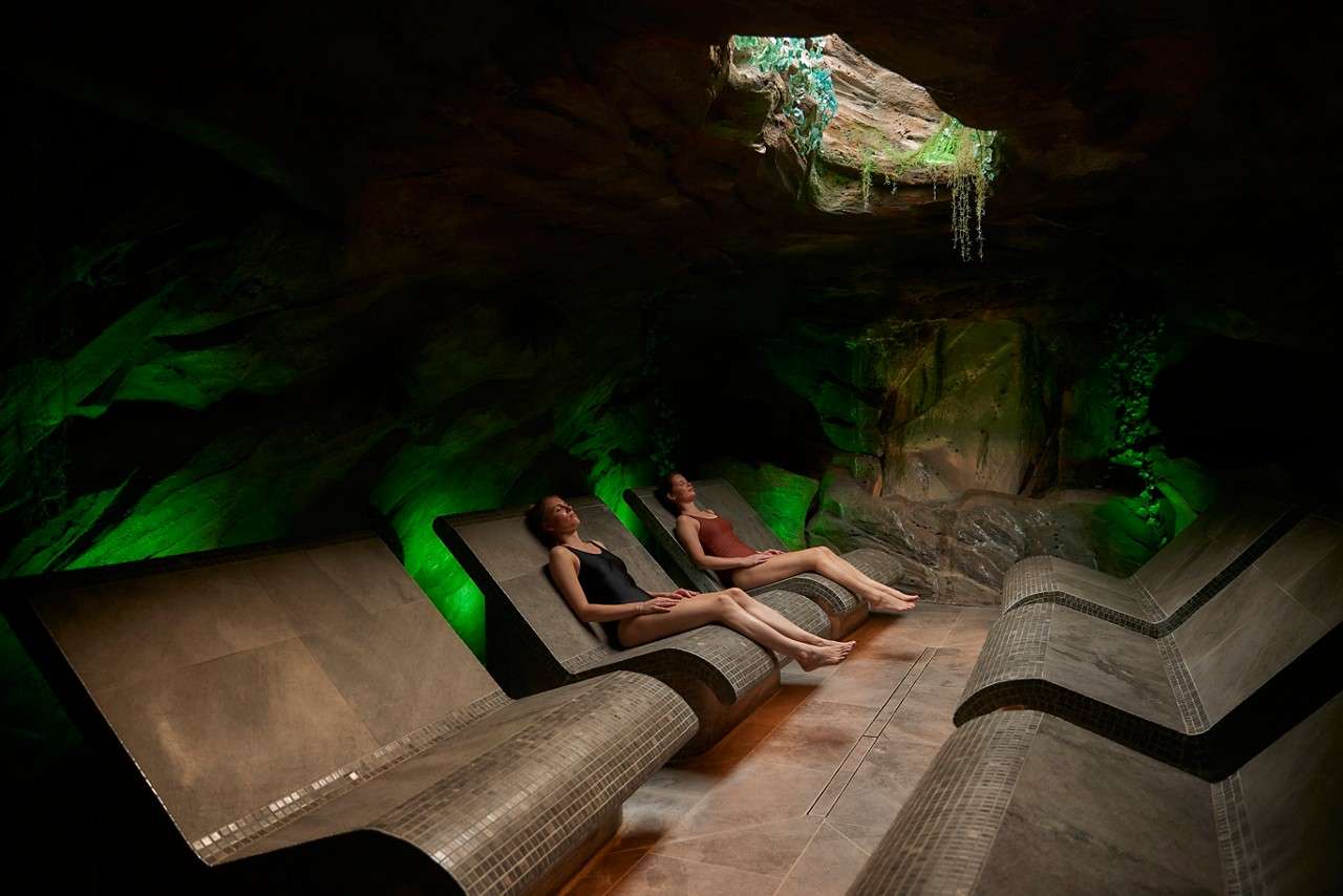 Two people recline on curved stone loungers, relaxing quietly, in a dim cave-like spa room with rock walls, green accent lighting, and a small skylight overhead with hanging plants.