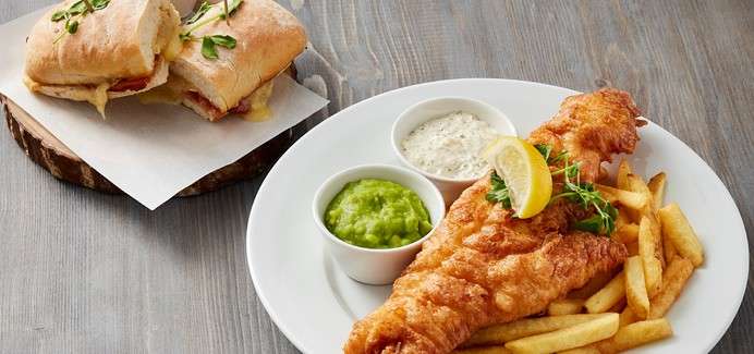 Golden fried fish is served beside fries, accompanied by mushy peas and tartar sauce, garnished with lemon and herbs; nearby sits a toasted sandwich on parchment atop a wooden board.