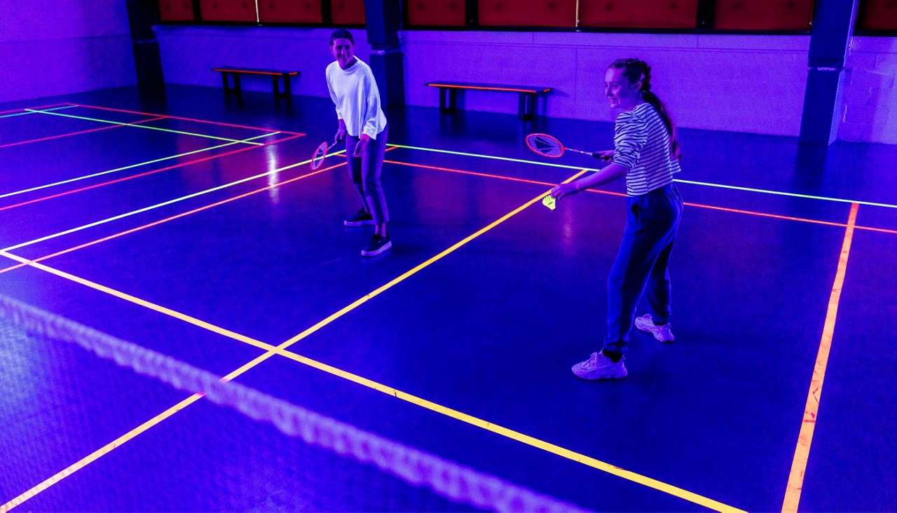 Two badminton players rally, holding racquets and a glowing shuttlecock, on an indoor court lit by UV lights; neon boundary lines, a foreground net, and benches along the back wall.