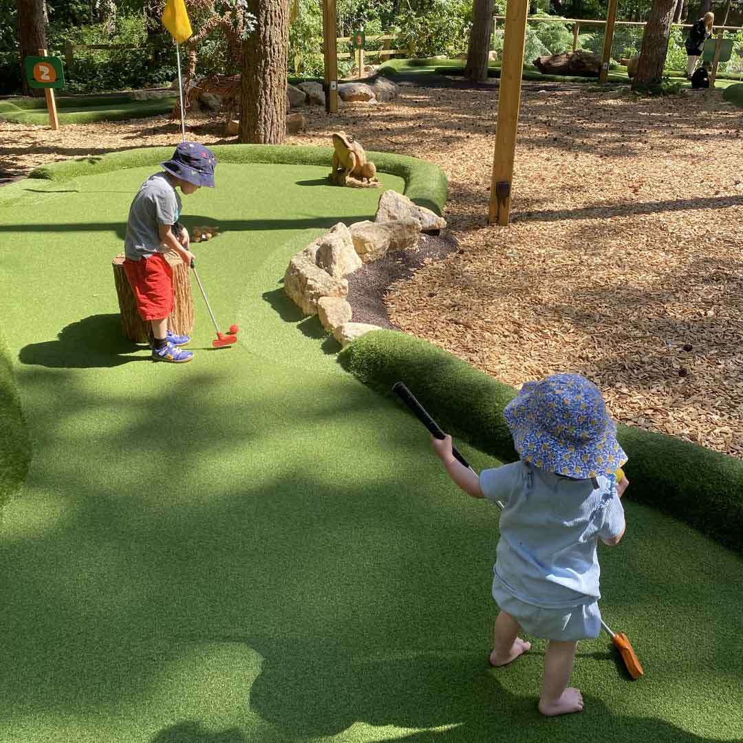 Two small children play mini-golf; one putts near a flag while another walks with a putter. Surrounded by artificial turf, trees, rocks, mulch, and a frog statue. Sign reads: “2”.