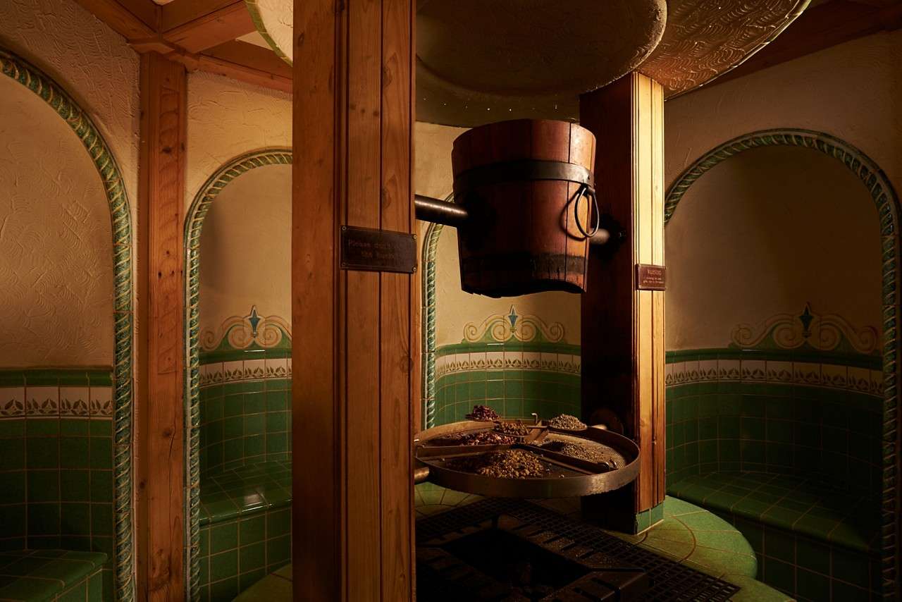 Wooden bucket hangs and drips water onto a circular tray of herbs, sitting between wooden posts, within a warmly lit, green-tiled steam room with curved benches and decorative arches.
