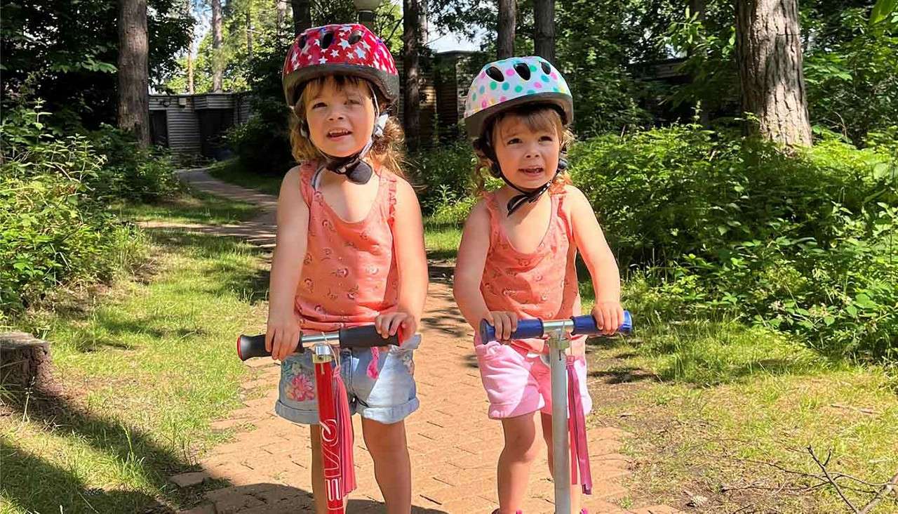 Two young children stand on kick scooters, wearing helmets, on a sunlit brick path in a leafy wooded park; one scooter displays the text "EVO".