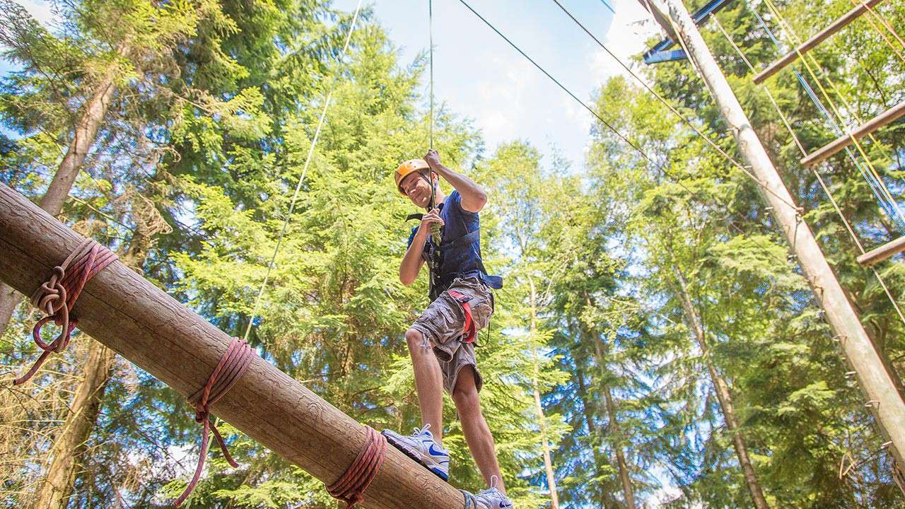 A harnessed person balances on a suspended log, gripping a safety rope. They wear a helmet and sneakers. The high-ropes course stands among tall evergreen trees under a bright sky.