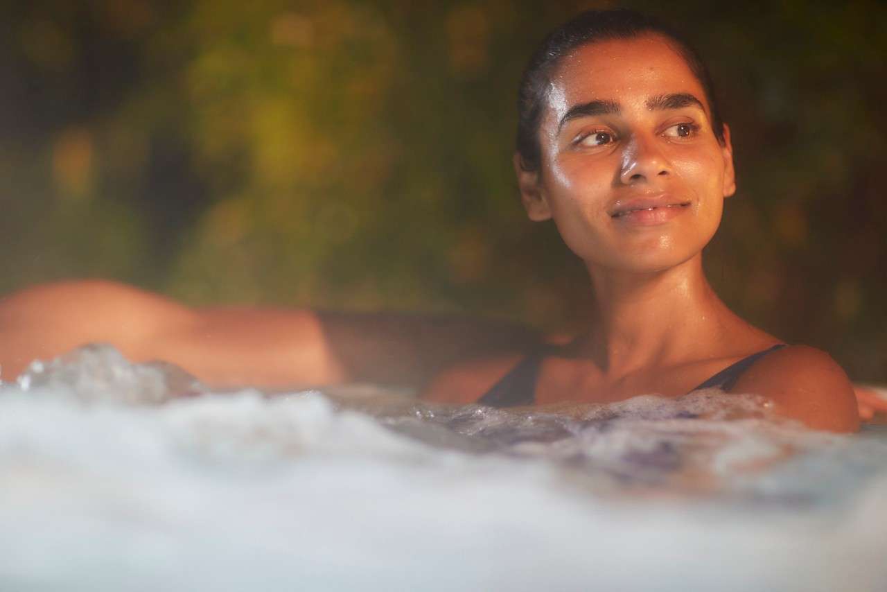 Person relaxing in a bubbling hot tub, smiling and looking to the side; warm light reflects on wet skin as foliage blurs in the evening background.