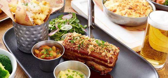 Glazed pork belly rests topped with crunchy crumbs, flanked by two sauces. Nearby, fries in a metal cup, leafy salad, a cheesy bake, and frothy beer on a wooden table.