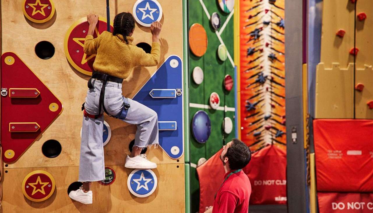 Climber ascends a colorful indoor wall using circular star holds; an instructor watches below in a padded arena with other themed routes. Red pads read: DO NOT CLIMB.