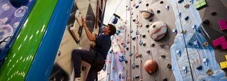 Climber ascends a vertical indoor wall, gripping plastic holds; belay ropes hang nearby. Context: colorful adjacent routes with planet-shaped volumes, bright panels, and various handholds across the rock-climbing gym.