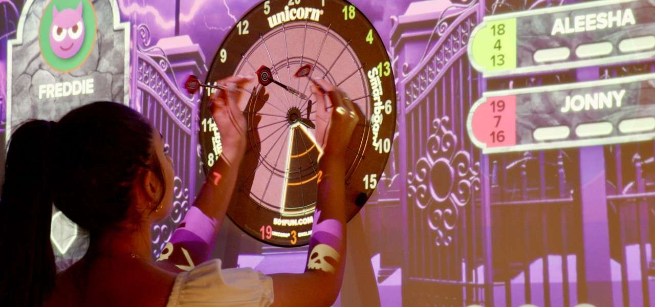 Player places darts on a projected electronic dartboard in a purple arcade setting with on-screen score panels. Text: FREDDIE; unicorn; Smartboard; ALEESHA; 18; 4; 13; JONNY; 19; 7; 16.