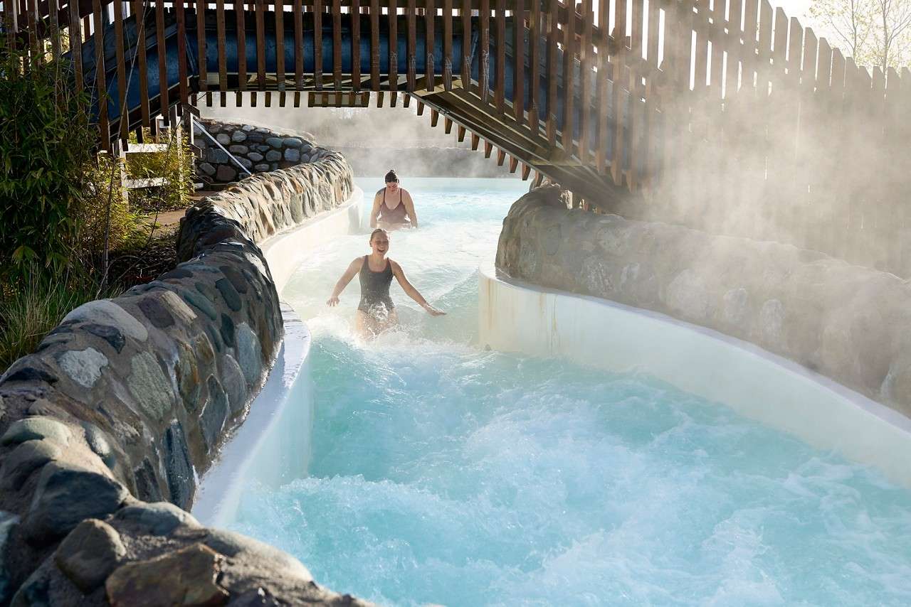 Two people splash down a twisting water slide, arms raised and laughing, passing beneath a small stone bridge amid rocky walls and surrounding greenery.