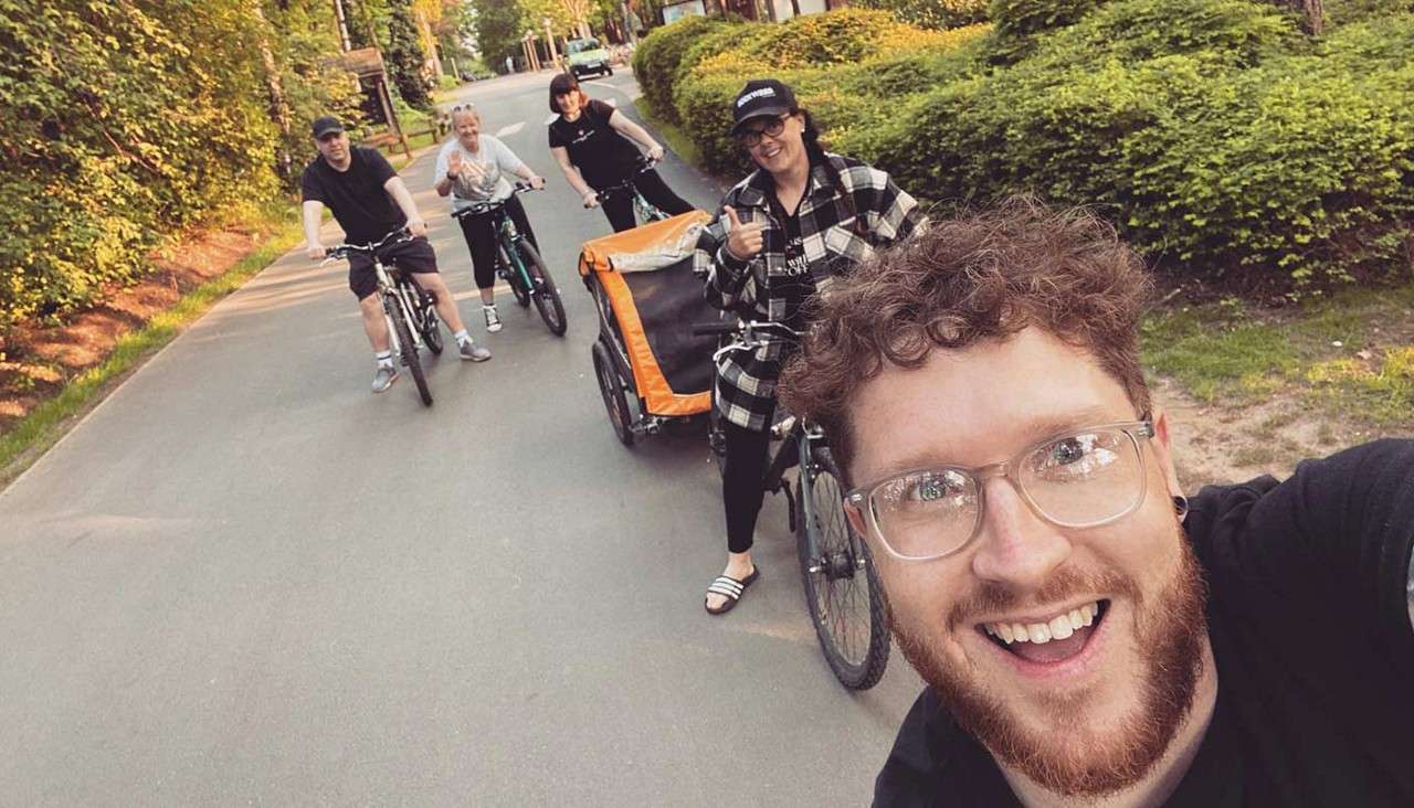 Smiling man takes a selfie while riding a bicycle; behind him, four friends cycle, one towing an orange child trailer, along a quiet, tree-lined suburban street in warm daylight.