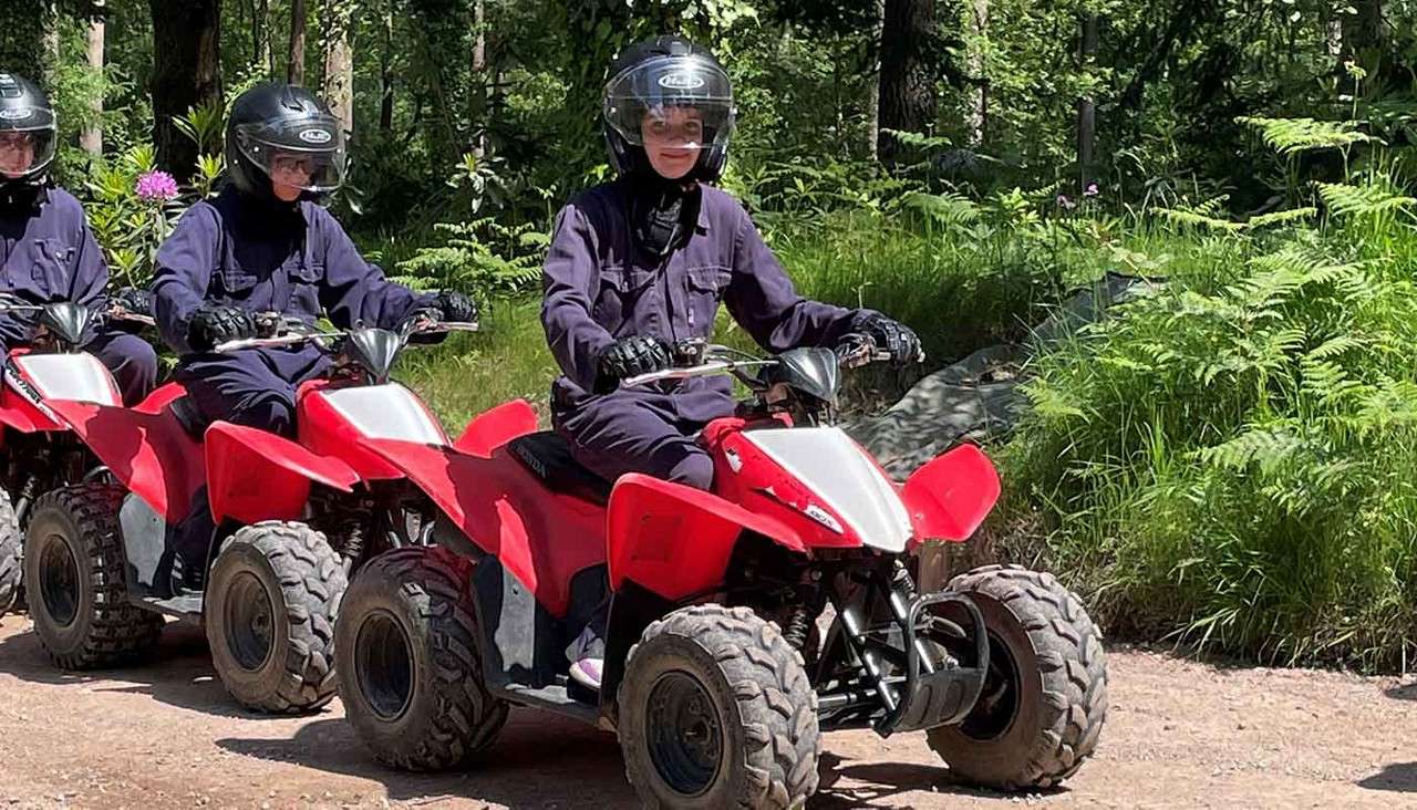 Riders drive red ATVs, wearing helmets and dark protective suits, moving in a line along a sunlit dirt trail through a green forest with ferns and trees.