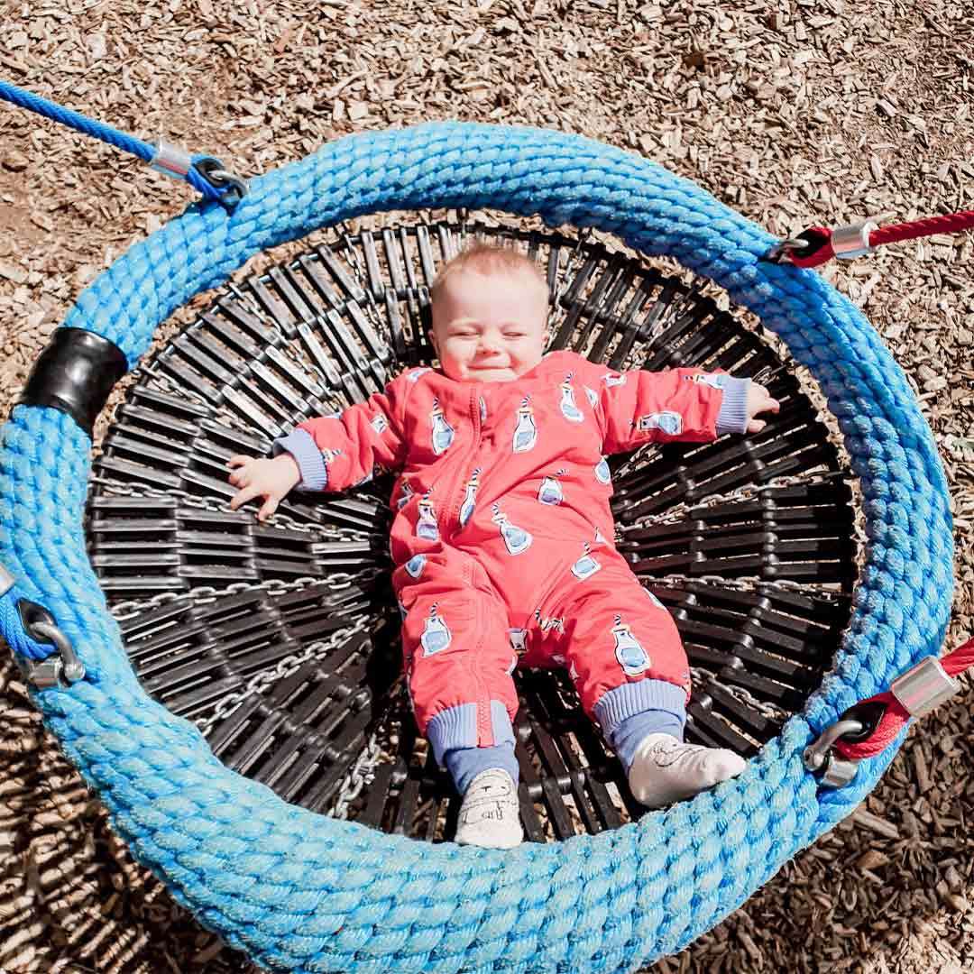Baby lies smiling on a round rope-net swing, arms spread, in a bright red patterned jumpsuit; swing suspended by blue ropes; playground ground covered with wood chips in sunlight.