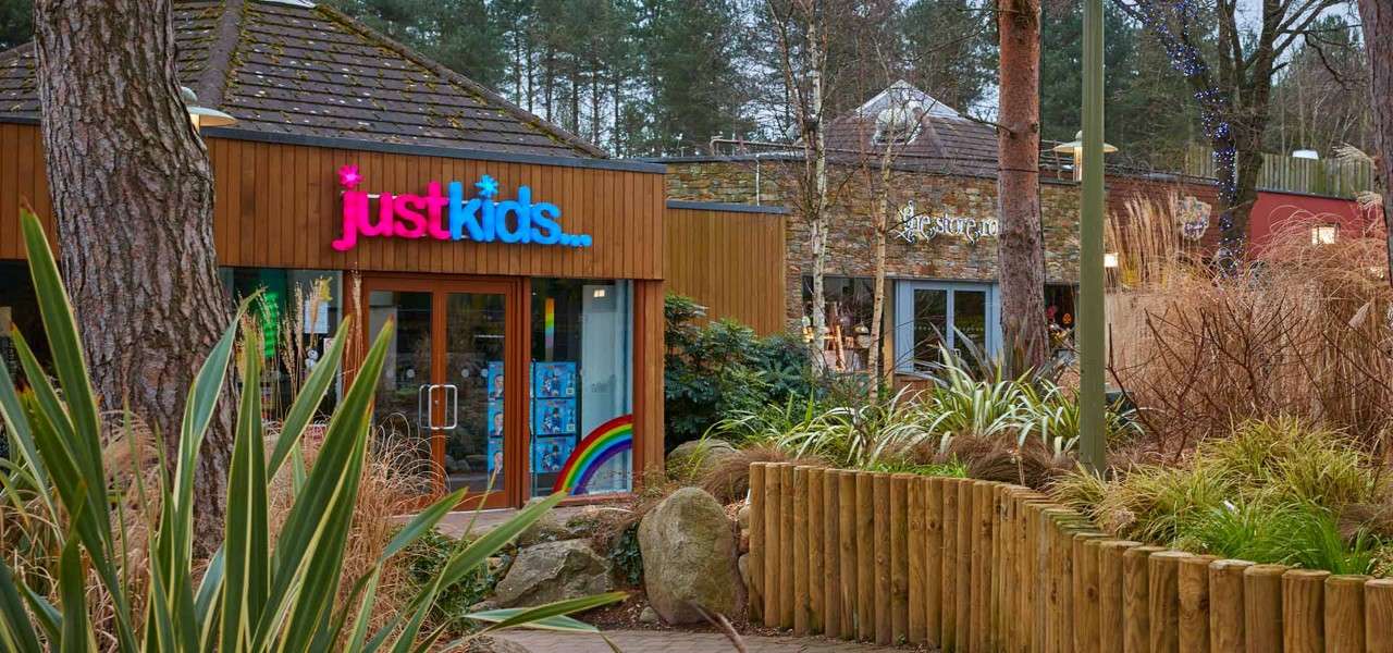 Toy store entrance displays sign “just kids...” and closed glass doors; neighboring building shows sign “the store room”; both sit among landscaped plants, wooden fencing, trees, and a forested background.