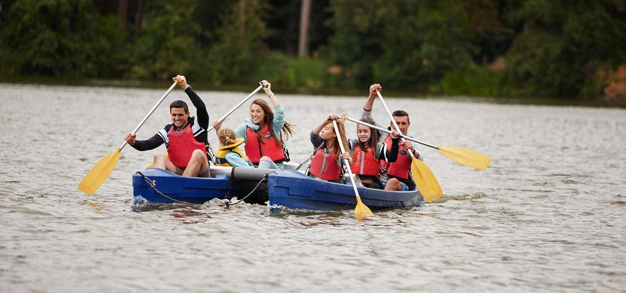 Group of people wearing red life vests paddle a twin-hulled blue canoe; a child sits between them; on a calm lake with a tree-lined shore.
