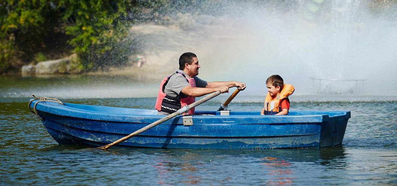 Blue rowboat carries an adult rowing and a child seated in life jackets, moving across a calm lake, with a water fountain mist and shoreline trees in the background.