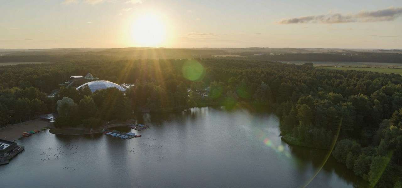 Sun sets, casting rays over a forested lakeshore; calm water reflects light while a dome-shaped building and small docks sit among trees, with distant fields and horizon under scattered clouds.