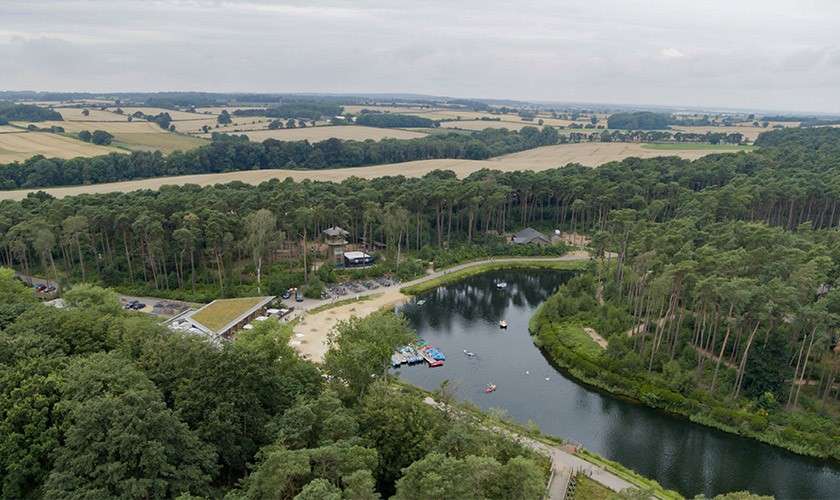 Lake hosts small paddleboats and a few moored craft, reflecting an overcast sky, while sandy beach, cabins, parking, and dense pine forest encircle it, with patchwork farmland stretching beyond.