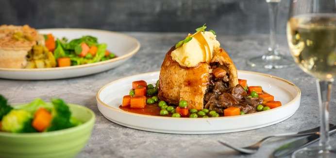 A dome-shaped pastry opens, revealing beef stew; mashed potato tops it. Gravy with peas and carrot cubes surrounds. Background shows side plates, a wine glass, and cutlery on a tabletop.