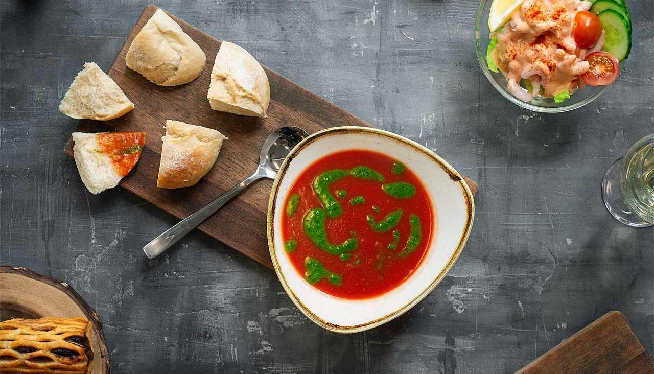Bowl of red soup sits garnished with green drizzle, accompanied by torn bread and spoon on wooden board; nearby, a shrimp salad, white wine, and pastry rest on gray tabletop.