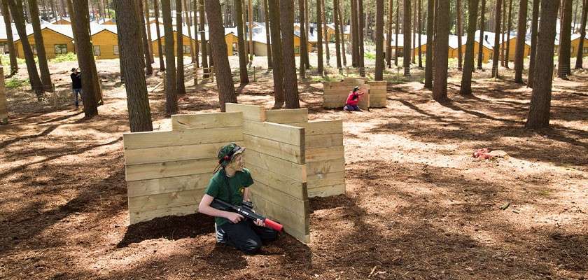 Players with laser-tag guns crouch and aim from behind wooden barricades, engaging in a mock skirmish. Tall pine trees surround them, with yellow cabins visible in the background across the forest floor.
