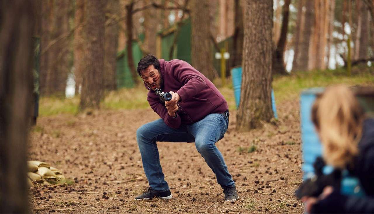 Player crouches and aims a paintball marker, bracing mid-stride; surrounding context is a wooded paintball field with trees, blue barricades, and another opponent blurred in the foreground.