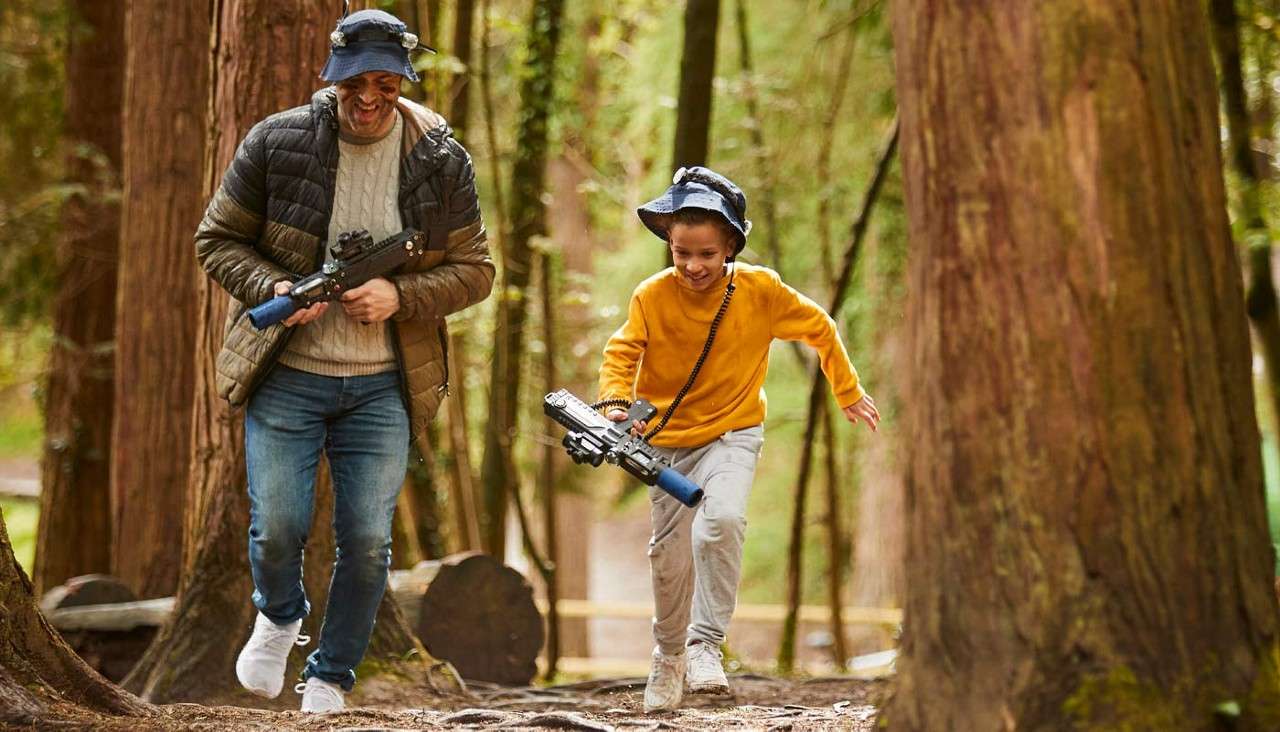 Two players run and laugh, carrying laser-tag blasters and wearing sensor hats, weaving between tall trees and logs along a forest trail.