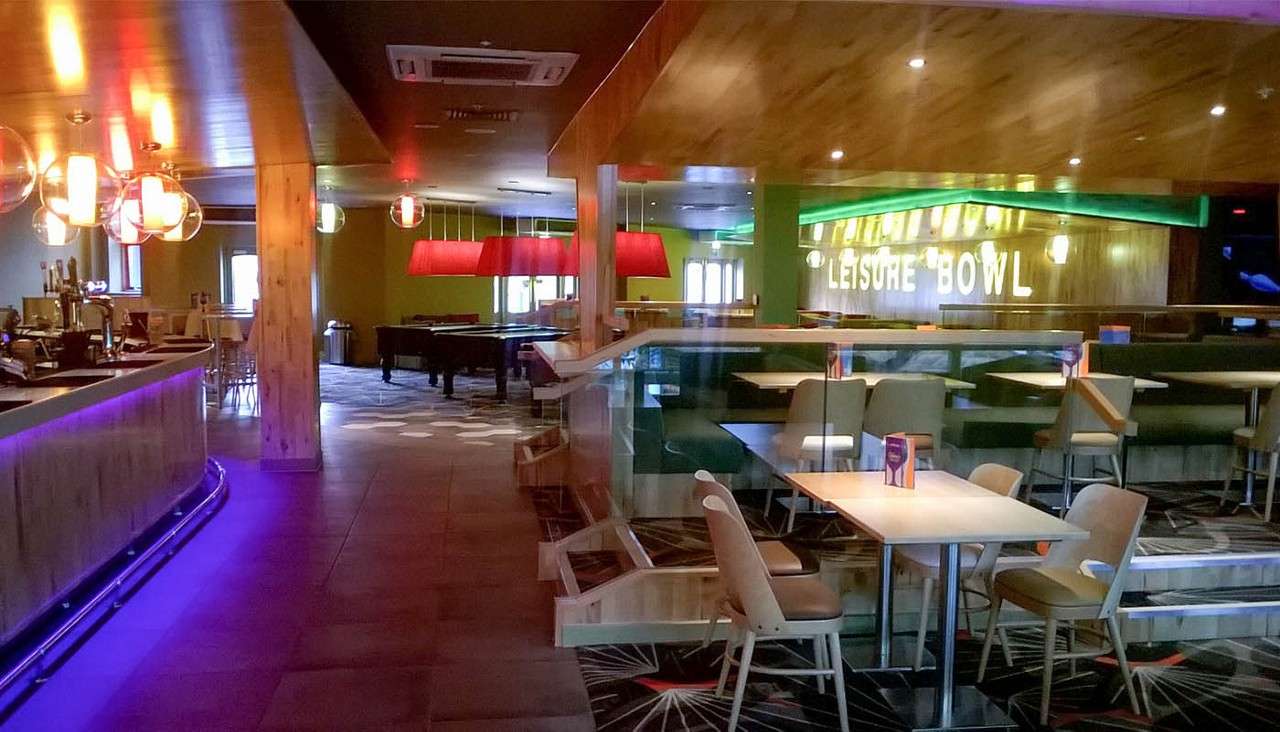 Bar and dining tables stand empty under purple and red lights; pool tables nearby. Context: modern leisure center with booths and a wall sign: LEISURE BOWL.