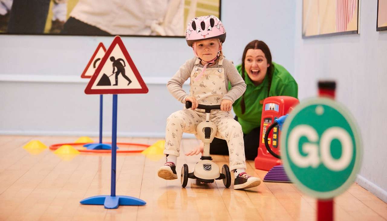 Child on a ride-on tricycle scoots indoors, wearing a pink helmet; cones and roadwork signs form a play course; a green sign reads GO, while an adult encourages behind.