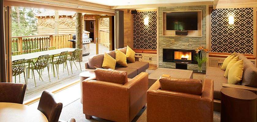 Living room seating clusters face a fireplace and wall-mounted TV, while sliding doors open to a wooded-deck dining area with green chairs and grill, blending indoor comfort with outdoor views.