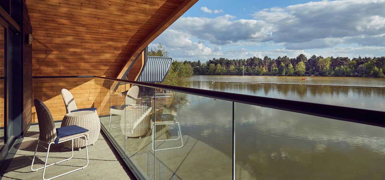 Wicker patio chairs sit on a curved wooden balcony, overlooking a calm lake bordered by trees under partly cloudy sky; glass railing frames the waterside view.