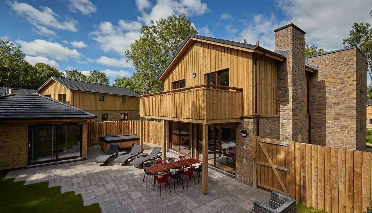 Wood-clad two-story house presents a balcony and sliding glass doors, overlooking a paved patio with dining table, loungers, and hot tub, enclosed by wooden fencing amid trees under blue sky.