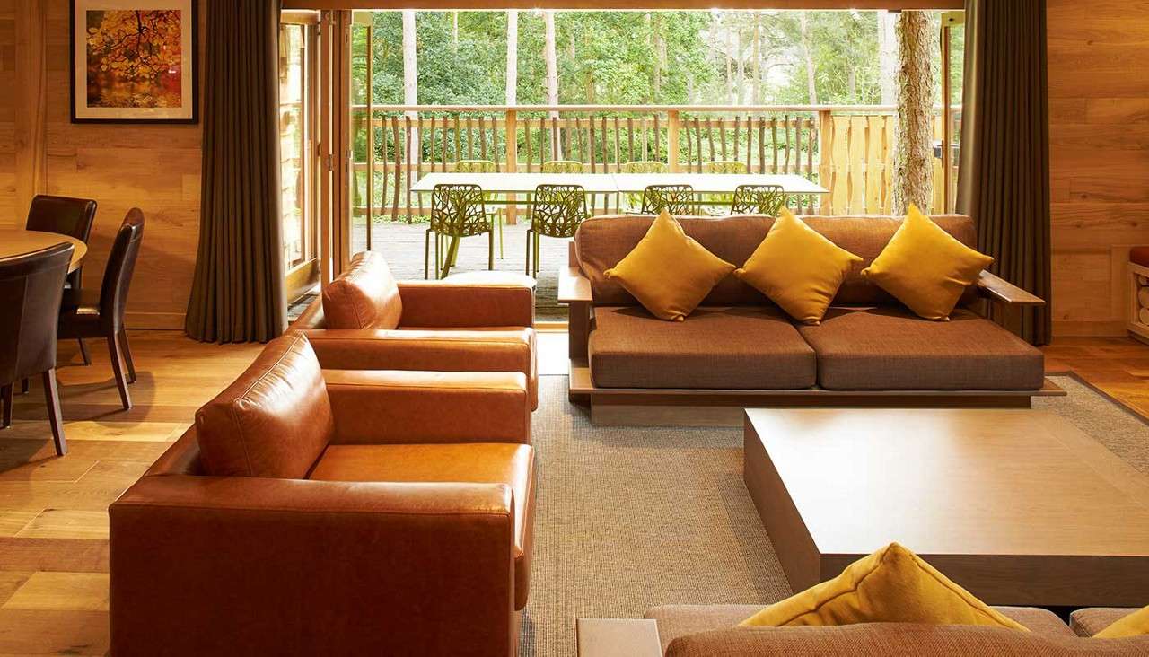 Brown sofa with yellow pillows faces low coffee table; leather armchairs flank it. Sliding doors open to patio with green chairs and long table, surrounded by forest; dining set left.