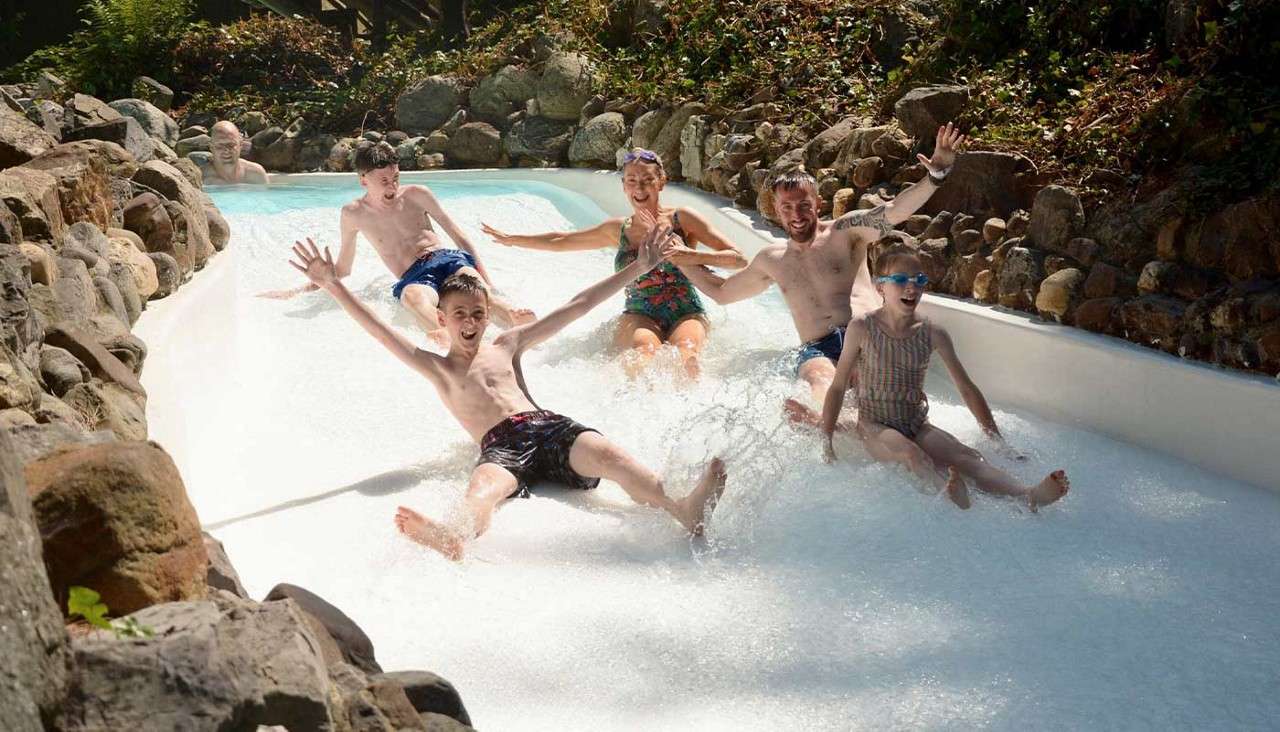 Group of six people slide down a shallow water flume, arms raised and splashing. Rock-lined channel curves through a sunlit outdoor area with foliage.