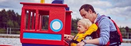 Adult and toddler steer a small red toy boat; both wear life jackets. Seated side by side, the adult guides the child’s hand, with water and trees in the background.