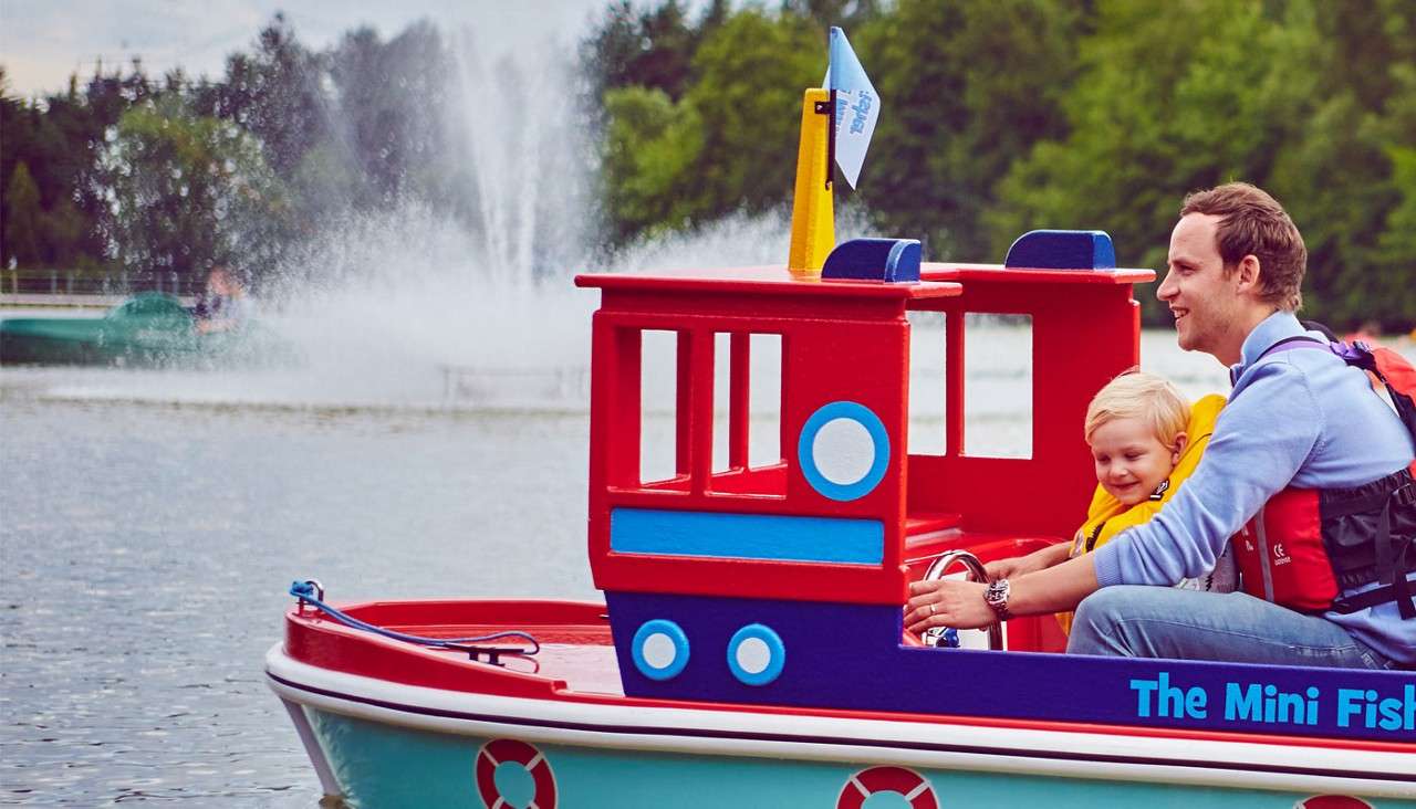 Toy-like red boat carries an adult and child wearing life jackets, cruising on a lake; fountains spray in the background, trees line the shore. Text: “The Mini Fis”.