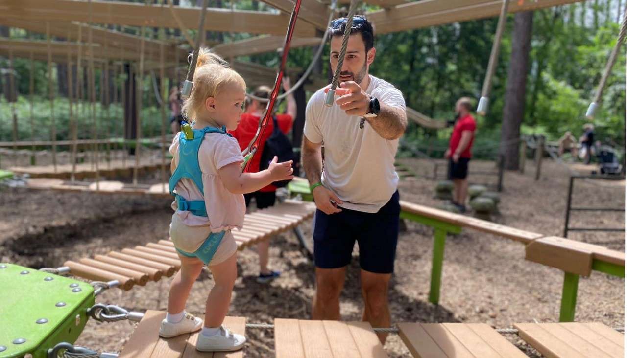 Toddler in a safety harness steps onto a suspended wooden bridge, while an adult man points and guides her, within a forest low-ropes adventure course.