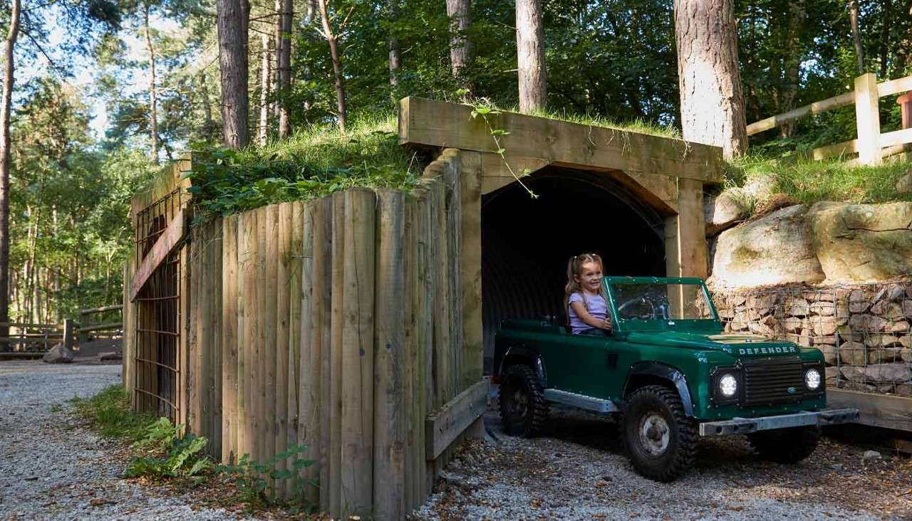 Miniature green off-road vehicle drives out of a wooden tunnel; a child steering, headlights on, gravel path beneath, surrounded by trees and fencing in a forest. Text on grille: DEFENDER.