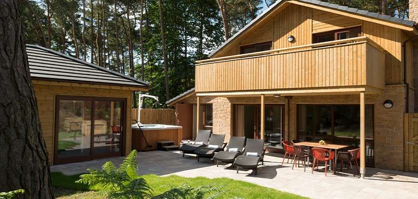 Wood-clad house features patio furnishings, with lounge chairs and dining table arranged for relaxation, beside a hot tub; set amid a forested yard, sunlight filtering through trees.