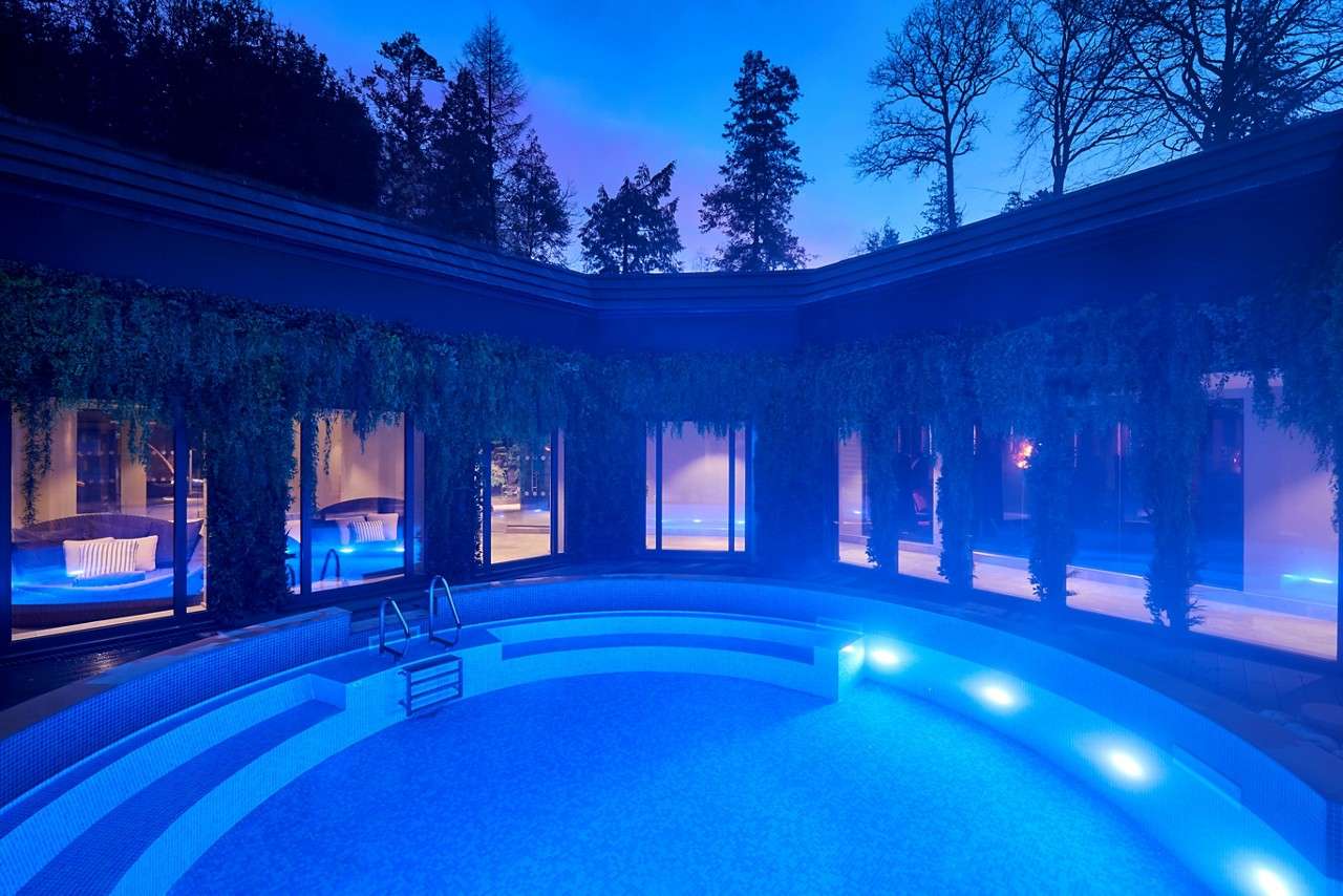 Curved swimming pool glows blue, steps and underwater lights illuminating tiers; surrounded by windows and hanging vines in a courtyard, with silhouetted trees against dusk sky.
