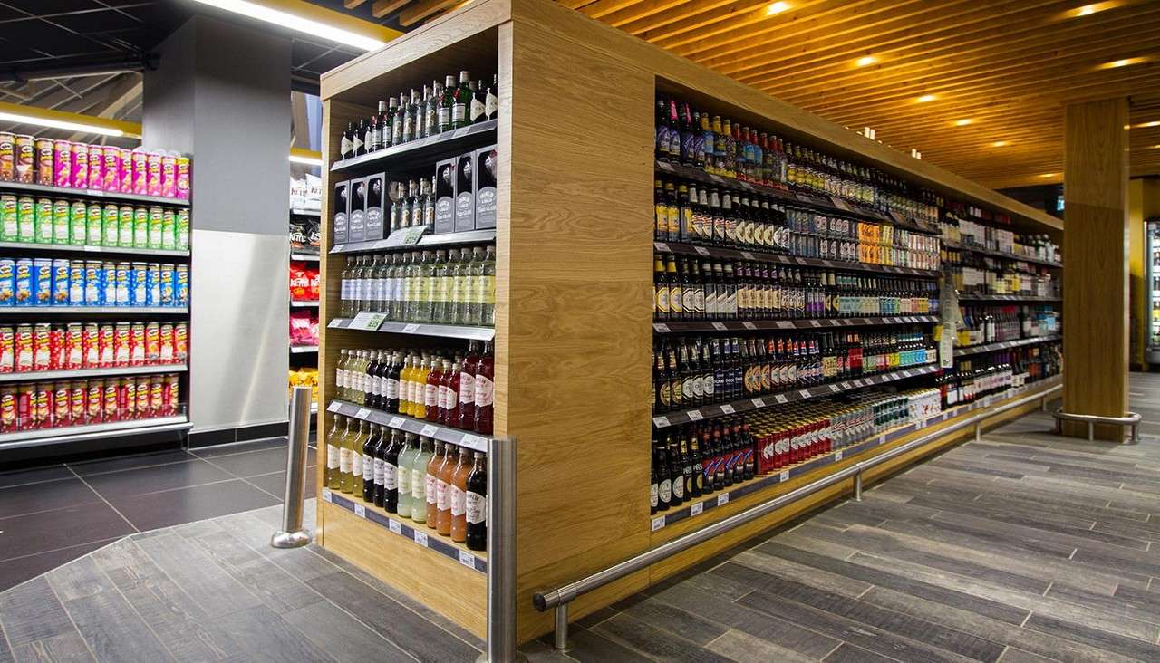 Shelving units display rows of bottled beverages and stacked snack cans, in long aisles of a brightly lit supermarket with wood ceiling and tile floors. Pringles.