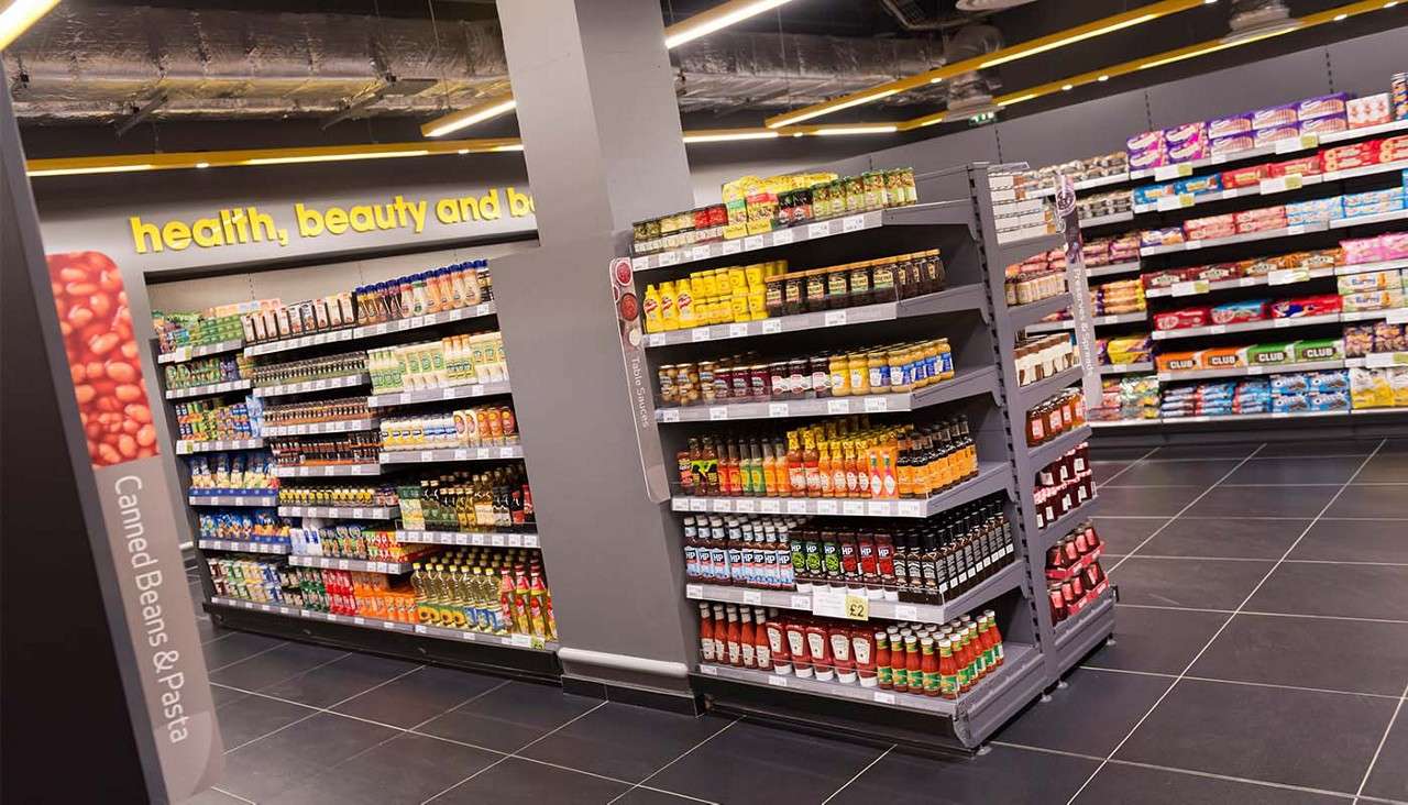 Shelves display sauces, condiments, drinks, and snacks, organized in a clean supermarket aisle with dark tile flooring, grey walls, and exposed ceiling ducts. Text: "Canned Beans & Pasta", "health, beauty and b", "£2", "HP", "CLUB".