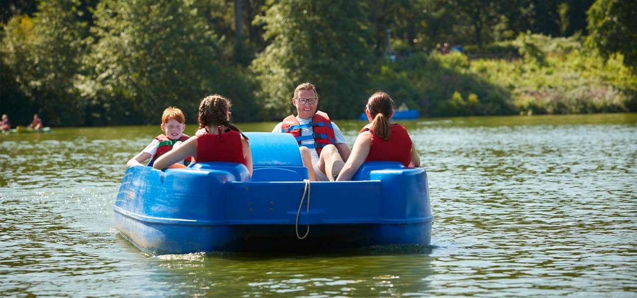 Four people on a blue pedal boat pedal and relax, wearing life jackets, while floating on a calm lake; wooded shoreline and other small boats appear in the sunny background.