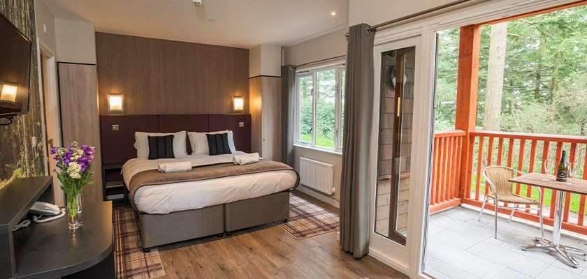 Bed stands neatly made; lamps glow. In context: modern hotel room with wood floors, desk with flowers, window and open sliding door to balcony with chairs, red railing, forest view.