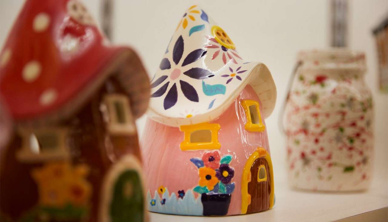 Colorful ceramic house figurine stands prominently, displaying a pink body, yellow-trimmed windows, flower-painted roof, and potted blooms; surrounded by other blurred decorative ceramics on a display shelf.