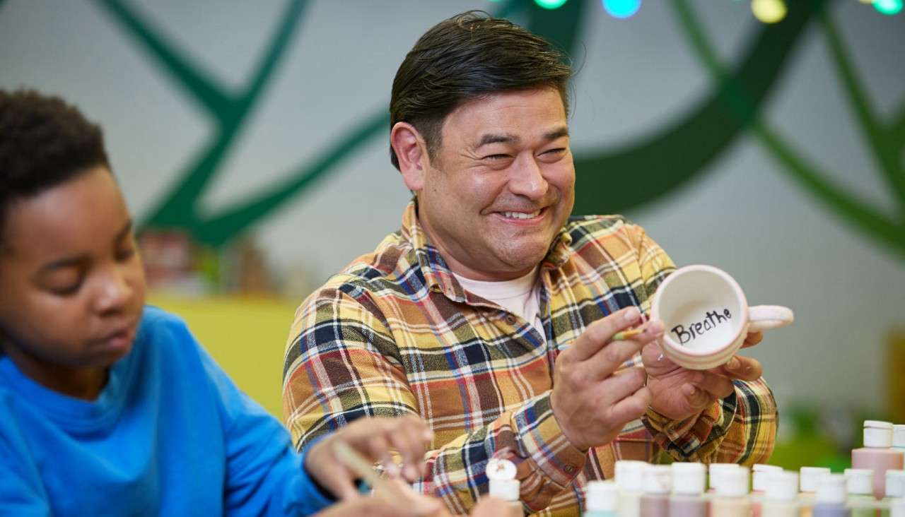Adult displays a ceramic mug, interior reading "Breath," while painting with a brush. Paint bottles cover the table, and another person works nearby in a colorful craft studio.