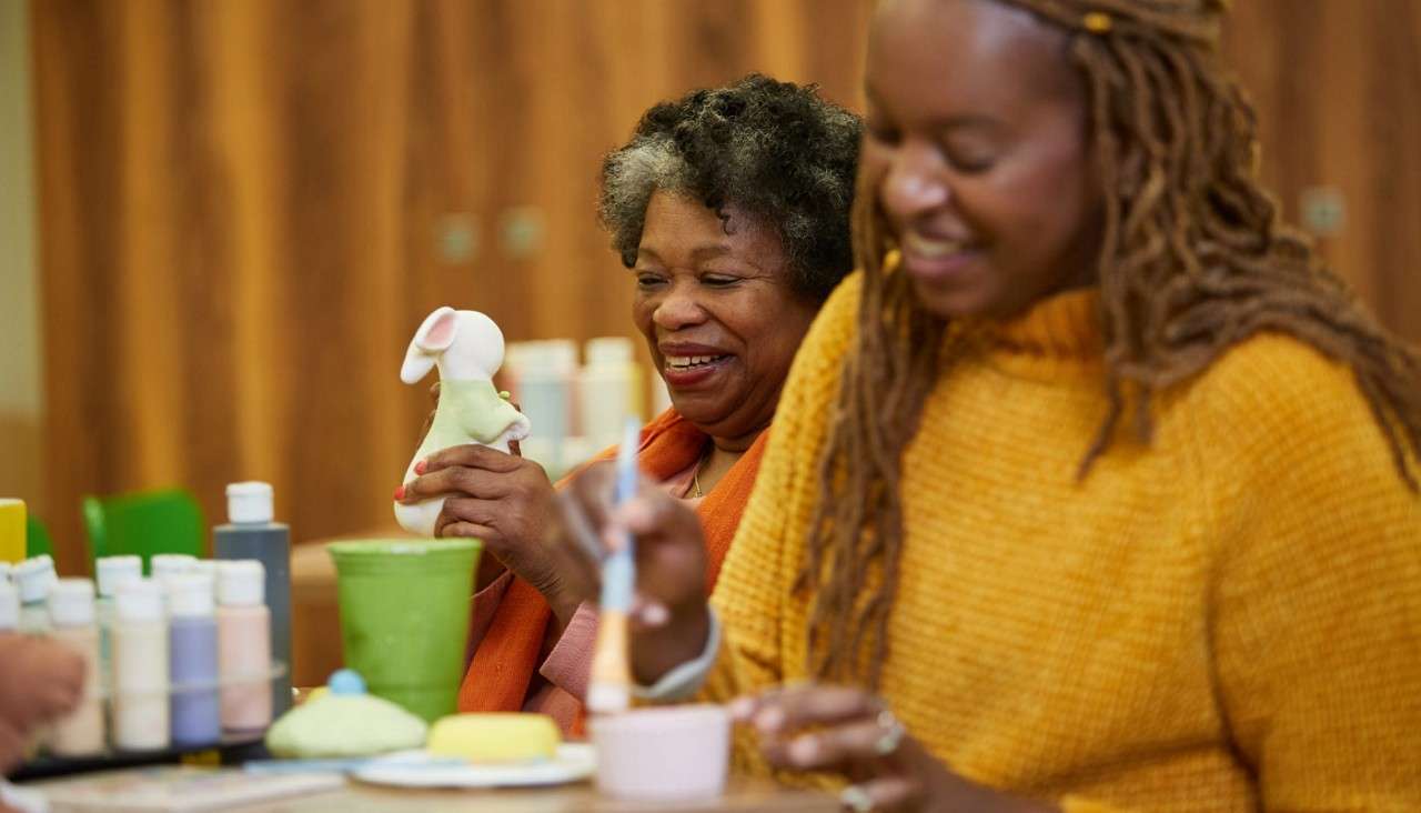Two people paint ceramics; one holds a small mouse figurine while the other brushes glaze on a cup. Bottles of paint sit on a table in a studio.